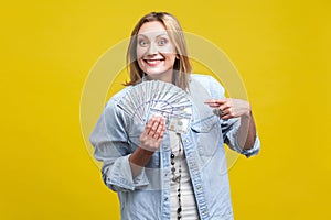 Portrait of happy lottery winner, pointing at fan of dollars, looking at camera with toothy smile.  on yellow background