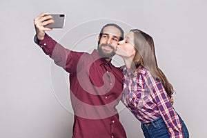 Portrait of happy in love satisfied couple standing and doing selfie, she is kissing her boyfriend and he is smiling at camera of