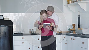 Portrait of happy loving couple hugging in kitchen and using tablet. Smiling young Caucasian man and woman spending