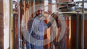 Portrait of happy male brewer admiring new equipment in brewery smiling walking at large beer tanks. Satisfied Caucasian