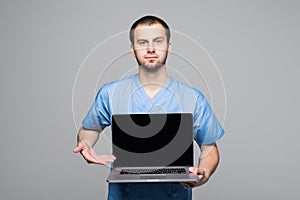 Portrait of a happy male doctor dressed in uniform with stethoscope showing blank screen laptop computer isolated over gray backgr