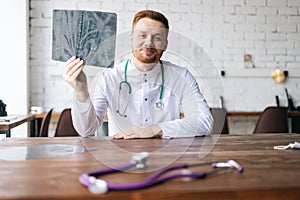 Portrait of happy male doctor in white uniform examining brain computerized tomography scan sitting at desk in hospital