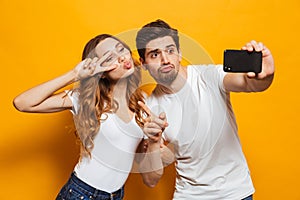 Portrait of happy man and woman taking selfie photo on smartphone and showing peace sign, isolated over yellow background