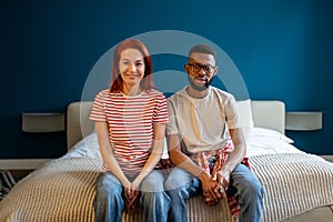 Portrait of happy married interracial diverse couple smiling at camera while sitting together on bed