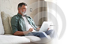 Portrait of happy mature man in casual clothes using laptop lying on sofa in house.