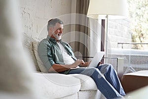 Portrait of happy mature man in casual clothes using laptop lying on sofa in house.