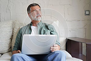 Portrait of happy mature man in casual clothes using laptop lying on sofa in house.