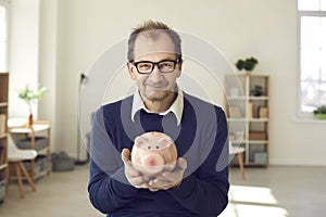Portrait of happy mature man in glasses holding piggy bank and smiling at camera