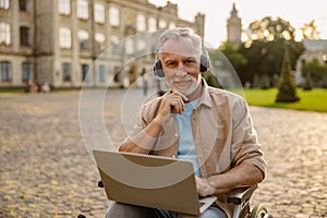 Portrait of happy mature man, recovering patient in wheelchair wearing headphones smiling at camera, using laptop