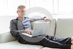 Portrait of happy mature man using laptop lying on sofa in house