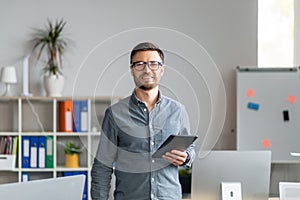 Portrait of happy mature office worker holding digital tablet, looking and smiling at camera, standing near workplace