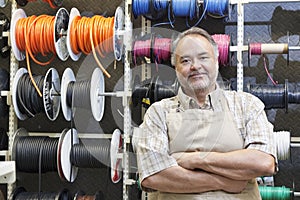 Portrait of a happy mature salesperson standing in front of electrical wire spool with arms crossed in hardware store