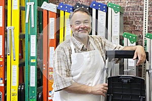 Portrait of happy mature store clerk standing by multicolored ladders in hardware shop