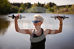 Portrait happy mature woman exercises with dumbbells outdoors, fitness training amidst beauty of nature, physical well