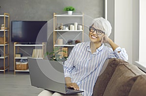 Portrait of happy mature woman sitting on sofa at home, holding laptop and smiling