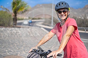 Portrait of happy mature woman, wearing protective helmet, on her bike in the sunny road. Healthy and active retirement concept