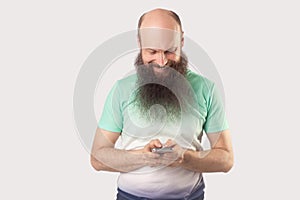 Portrait of happy middle aged bald bearded man standing, holding mobile smart phone and looking at screen, reading something with
