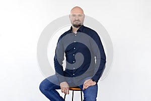 Portrait of happy middle-aged bald bearded man wearing blue shirt and jeans, sitting on stool on white background.