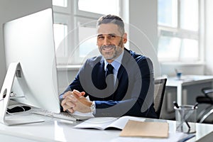Portrait of happy middle aged businessman sitting in front of computer at workplace in office, smiling to camera