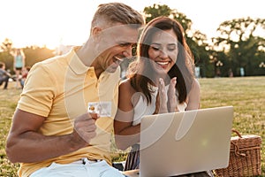 Portrait of happy middle-aged couple holding credit card while using laptop computer during picnic in summer park