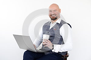 Portrait of happy middle-aged man in grey vest, blue jeans, white shirt, sit holding laptop and takeaway cup of coffee.