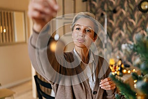 Portrait of happy middle-aged woman holding gold ball for decoration Christmas tree at home, closeup, bokeh lights