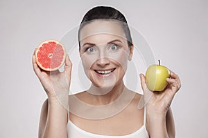 Portrait of happy middle aged woman with perfect face skin toothy smiling, holding fresh slice grapefruit and apple in her hands,