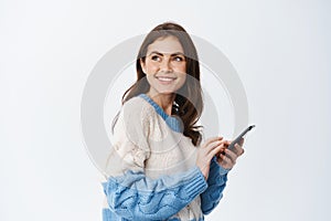 Portrait of happy milennial woman chatting in app, holding smartphone and turn back at logo on copy space, standing in