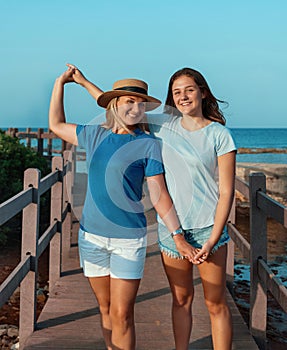 Portrait of happy mother and teenage daughter standing on pier at sea coast, holding hands and smiling, t-shirts mockup