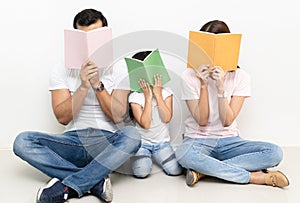 Portrait of happy multiethnic family sitting on the floor hiding behind colorful book at home. Family and childhood concept