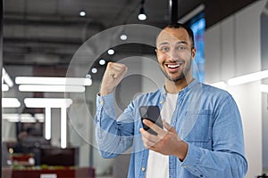 Portrait of a happy Muslim man standing in a modern office, holding a phone in his hand and rejoicing in success and