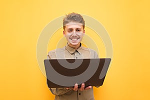 Portrait of a happy nerd in glasses and shirt on a yellow background, holds a laptop in his hand and looks into the camera with a