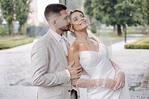 Portrait happy newlyweds walking outdoors. Handsome groom in light colour suit and bride in elegant wedding dress