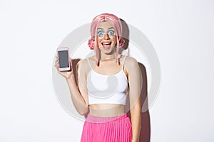 Portrait of happy party girl with pink wig and bright makeup, showing mobile phone screen with amused face, demonstrate