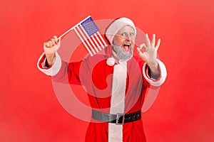 Portrait of happy patriotic elderly man with gray beard wearing santa claus costume holding USA flag