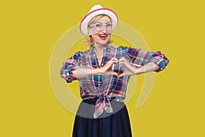 Portrait of happy peaceful modern stylish mature woman in casual style with white hat standing with hand heart shape gesture,