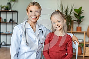Portrait of happy pediatrician and pretty child girl looking and smiling at camera, sitting in doctor's office