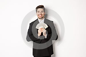 Portrait of happy and pleased handsome man in suit, hugging money and looking satisfied, standing over white background