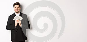 Portrait of happy and pleased handsome man in suit, hugging money and looking satisfied, standing over white background