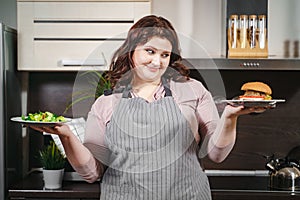 Portrait of a happy plus size woman holding a plate with burger and plate and salad in the kitchen. The concept of choosing