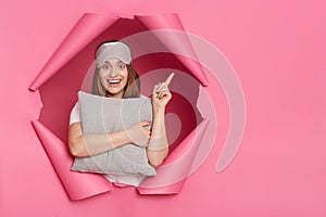 Portrait of happy positive Caucasian young adult woman with pillow in hands breaks through pink paper background, pointing finger