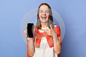 Portrait of happy positive woman in white T-shirt and orange sweater tied over shoulders, showing smart phone with empty display,