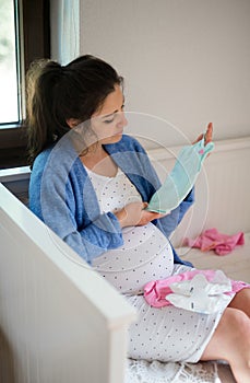 Portrait of happy pregnant woman indoors at home, holding baby clothes.