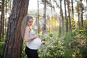 Portrait of happy pregnant woman outdoors in nature, touching her belly.