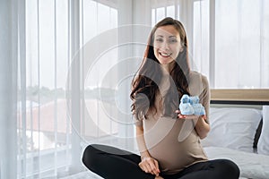 Portrait of happy pregnant woman sitting on the bed and holding small shoes for the unborn baby in white bedroom