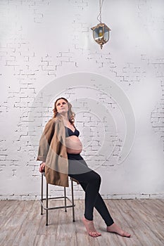 Portrait of a happy pregnant woman sitting on a chair in full growth against a background of a white brick wall. Smiling adult