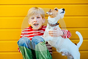 Portrait of happy preteen boy sitting on the ground playing with his yawning dog Jack Russell Terrier on yellow background outdoor