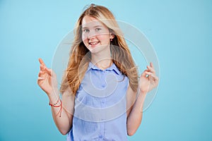 Portrait of happy pretty teenage girl with long fair hair wearing blue shirt, raising hands, dancing on blue background.