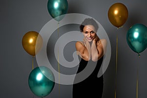 Portrait of happy pretty woman in evening black dress smiling with beautiful toothy smile posing against air balloons on gray wall