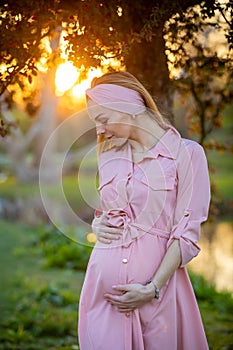 Portrait of a happy and proud pregnant young woman looking at her belly in park at sunrise with a warm back light in the